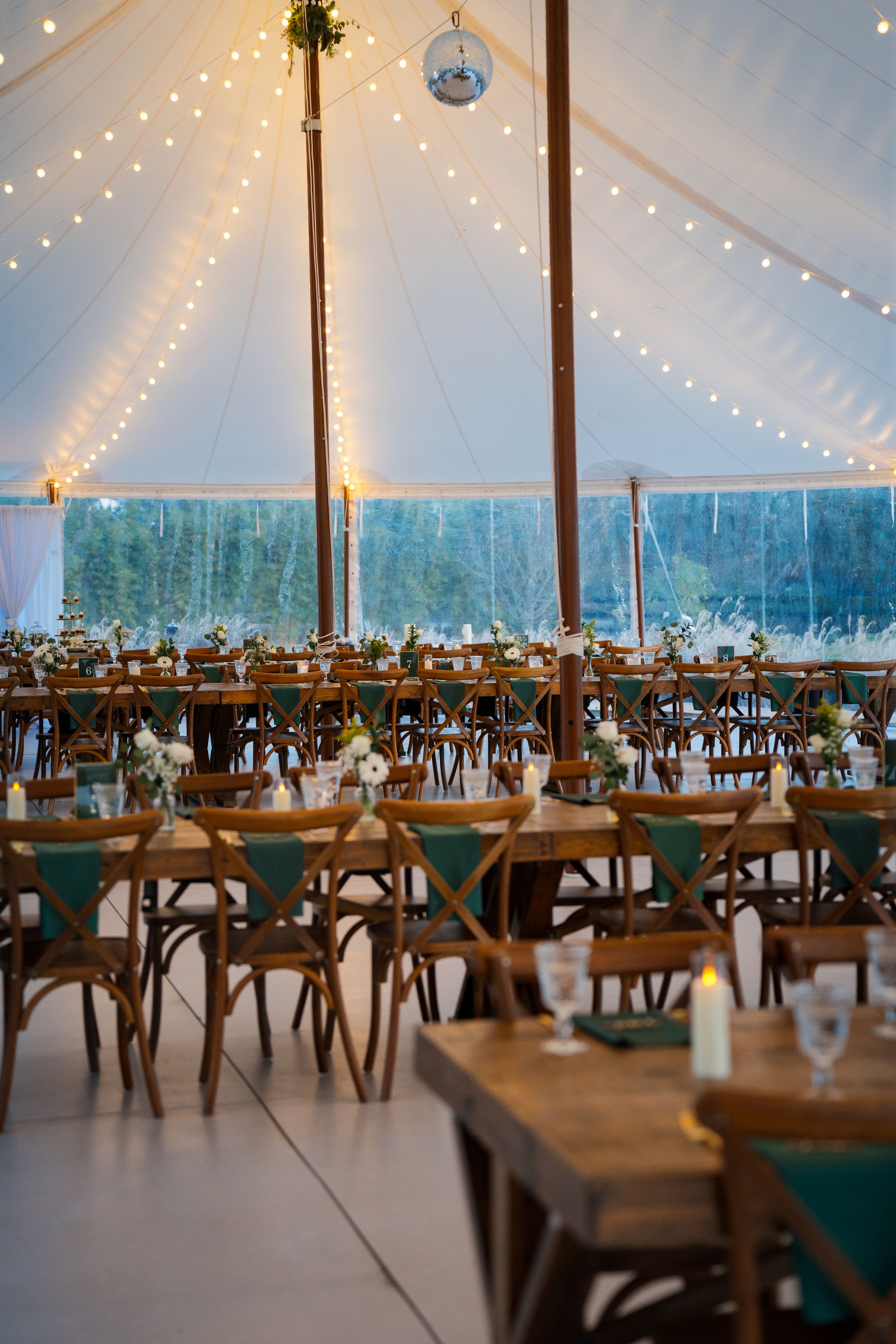 Elegant wedding reception tent with string lights, wooden cross-back chairs and rustic table settings under white canopy.