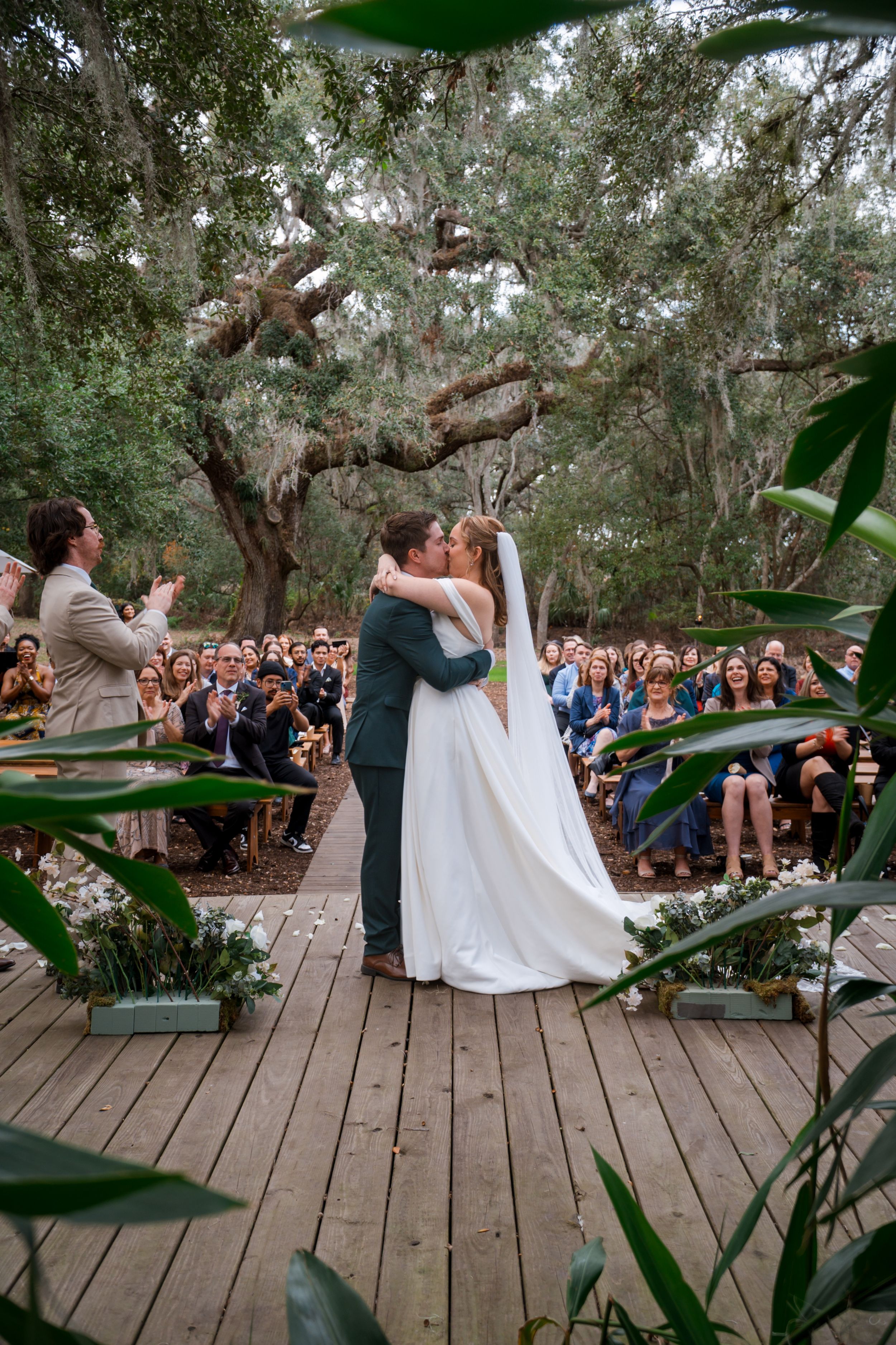 A beautiful outdoor wedding ceremony takes place on a wooden deck surrounded by trees and lush greenery.