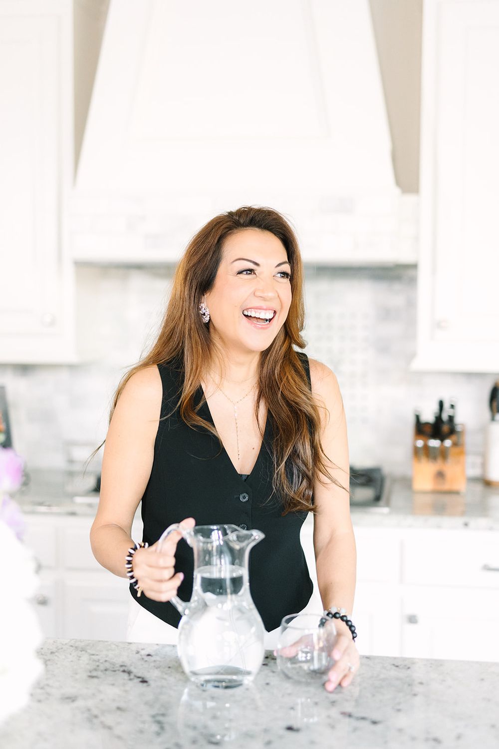 Woman in black sleeveless top laughs while standing at marble kitchen counter with glass water pitcher.