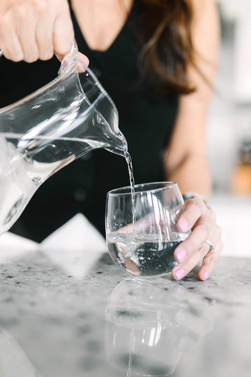 Water being poured from a clear glass pitcher into a drinking glass on a marble countertop.