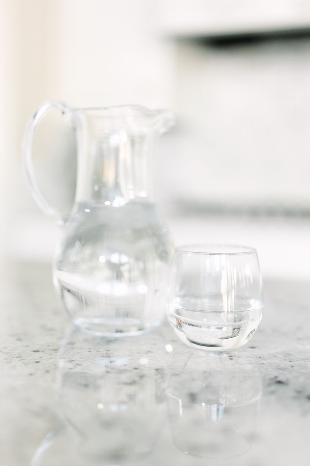 A clear glass pitcher and drinking glass with water on a light surface with soft, bright lighting.