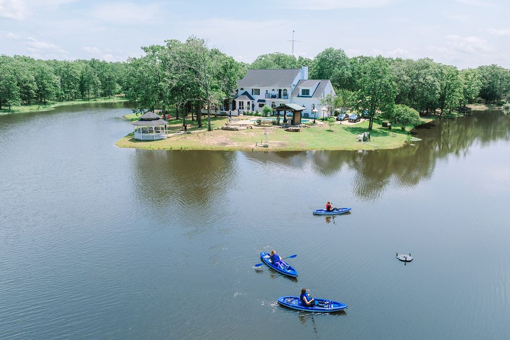 Aerial view of lakeside house with kayakers paddling on calm waters surrounded by green trees.
