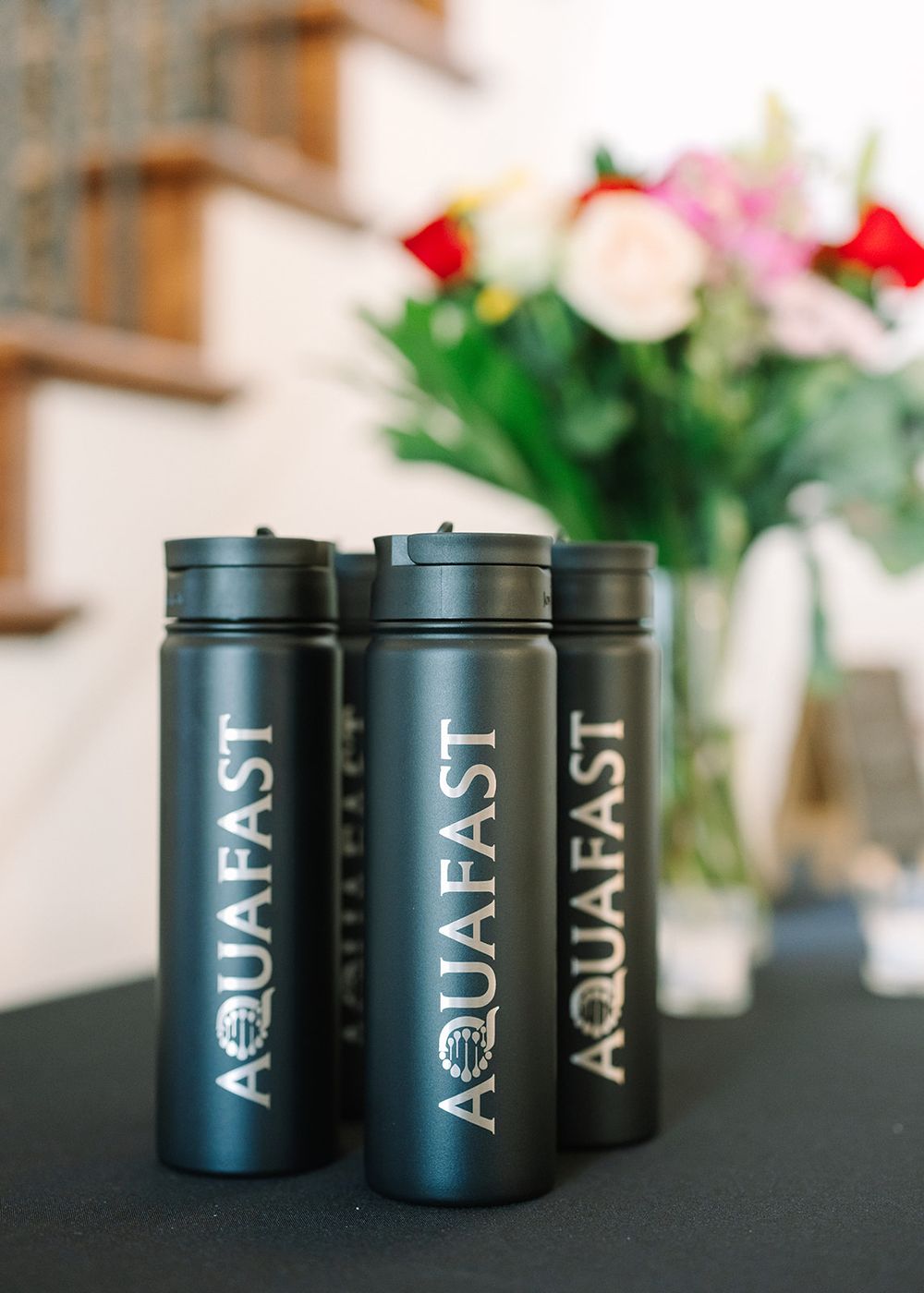 Three black Aquafast reusable water bottles displayed on a dark surface with flowers in the background.