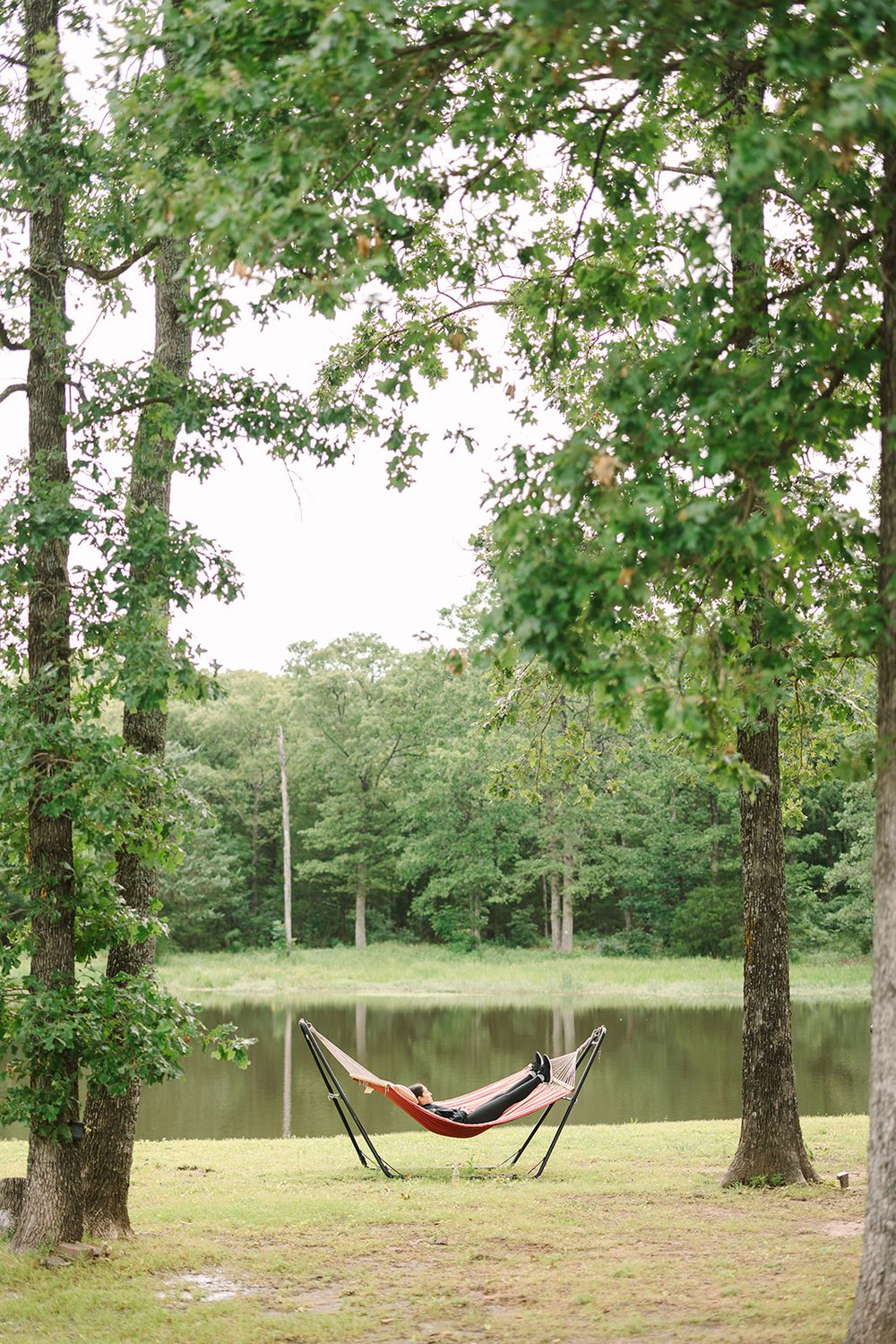 A red hammock stands on a portable frame between two trees near a serene lake in a peaceful woodland setting.