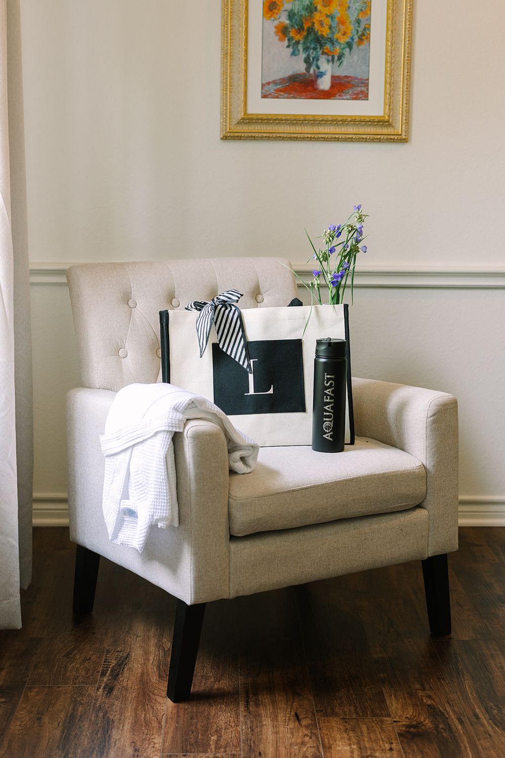 A cream-colored tufted armchair with shopping bags and flowers sits against a wall with framed artwork above.