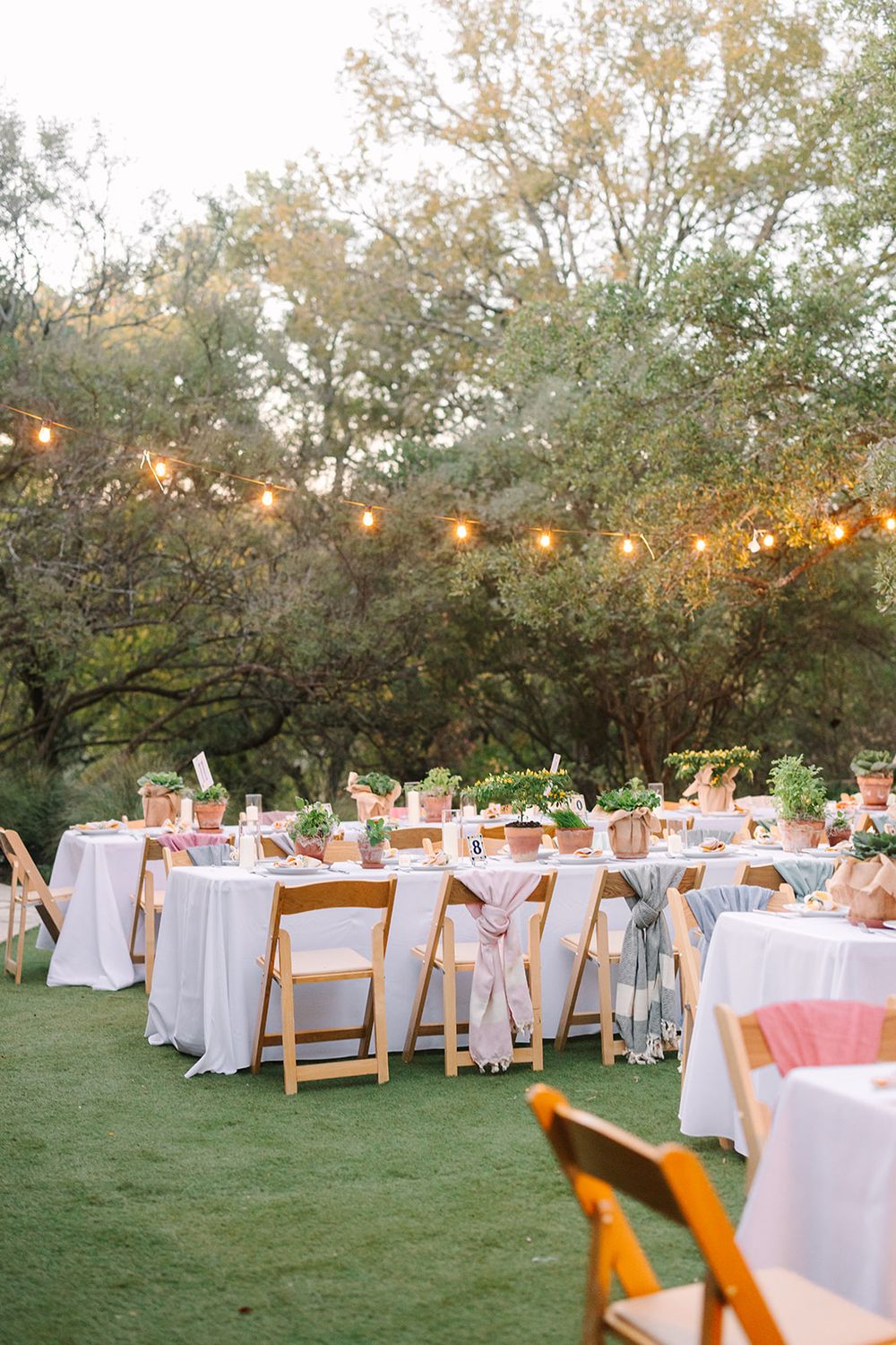 An outdoor wedding reception setup with white-clothed tables and string lights hanging from trees at dusk.