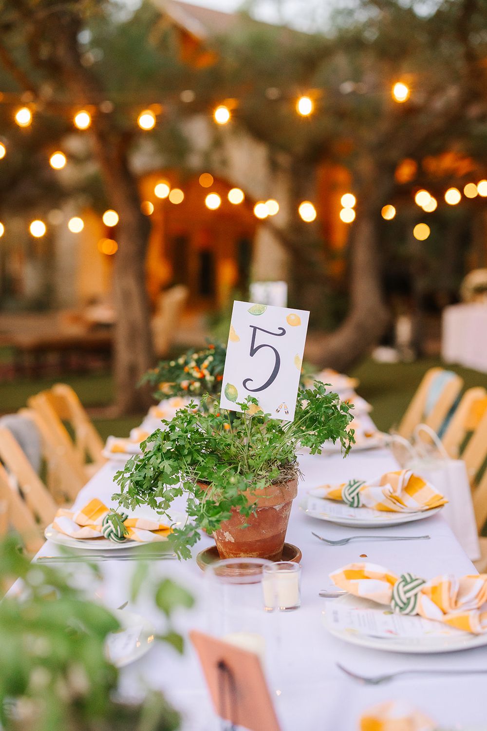 Elegant outdoor reception dinner table settings with string lights and table numbers decorated with greenery at dusk.