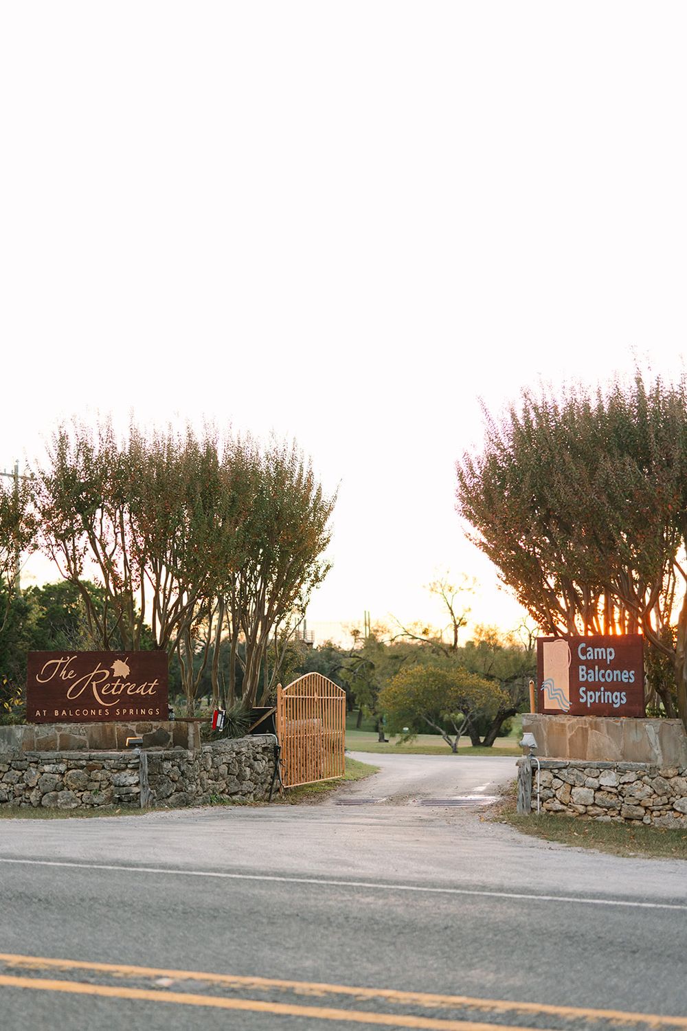 Stone entrance with signage for a vineyard estate with tall trees lining a curved driveway at sunset.