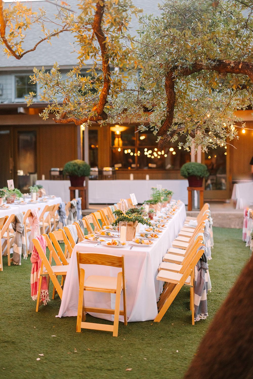 Elegant outdoor wedding reception setup with long tables, string lights, and wooden chairs under trees at dusk.