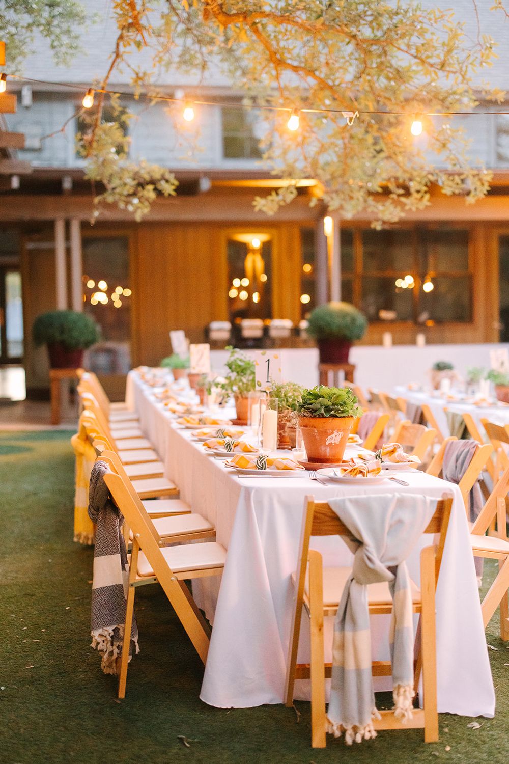 Elegant outdoor dining table set with white linens and terracotta pots under string lights at dusk for a garden party.