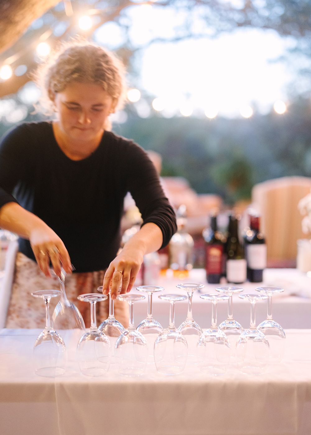 Person arranging delicate wine glasses and table settings for an elegant outdoor dining event or wine tasting setup.
