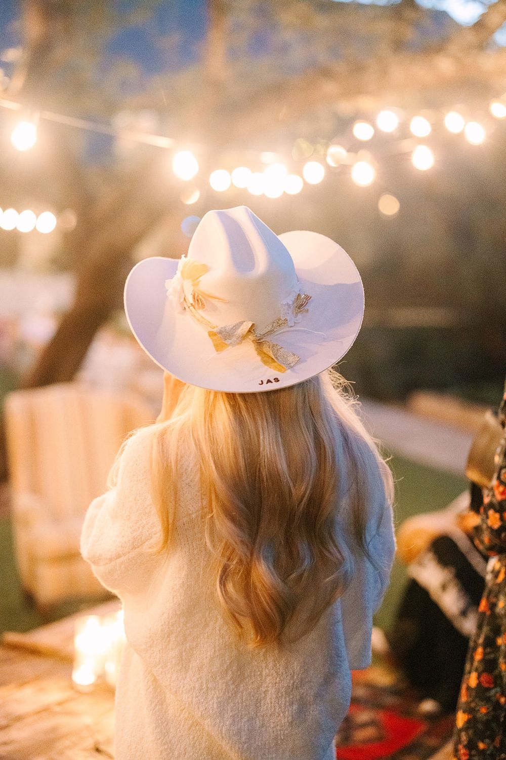 Person wearing white hat and sweater at outdoor event with string lights creating warm bokeh effect in background.