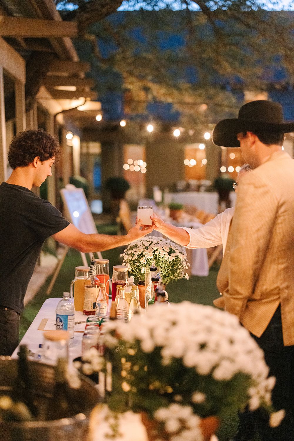 A bartender serves drinks at an outdoor evening reception decorated with floral arrangements and string lights.