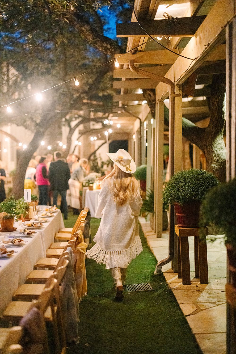 Evening outdoor dinner party under string lights with long dining table and person in white dress and cowboy hat walking.