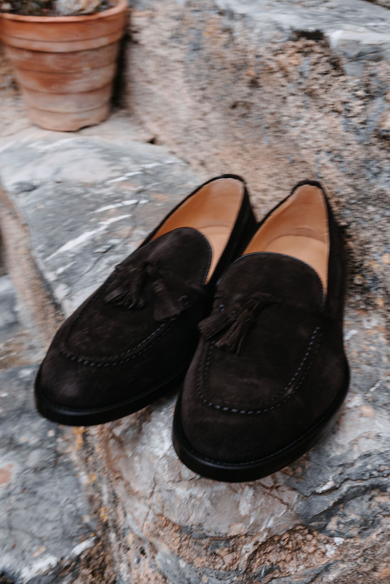 Ein Paar klassische dunkelbraune Wildleder-Loafer mit Quasten auf einer strukturierten Steinoberfläche mit Terrakottatöpfen im Hintergrund.