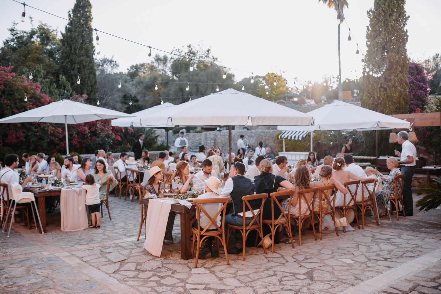 Outdoor wedding reception on stone patio with white umbrellas and wooden cross-back chairs during golden hour.