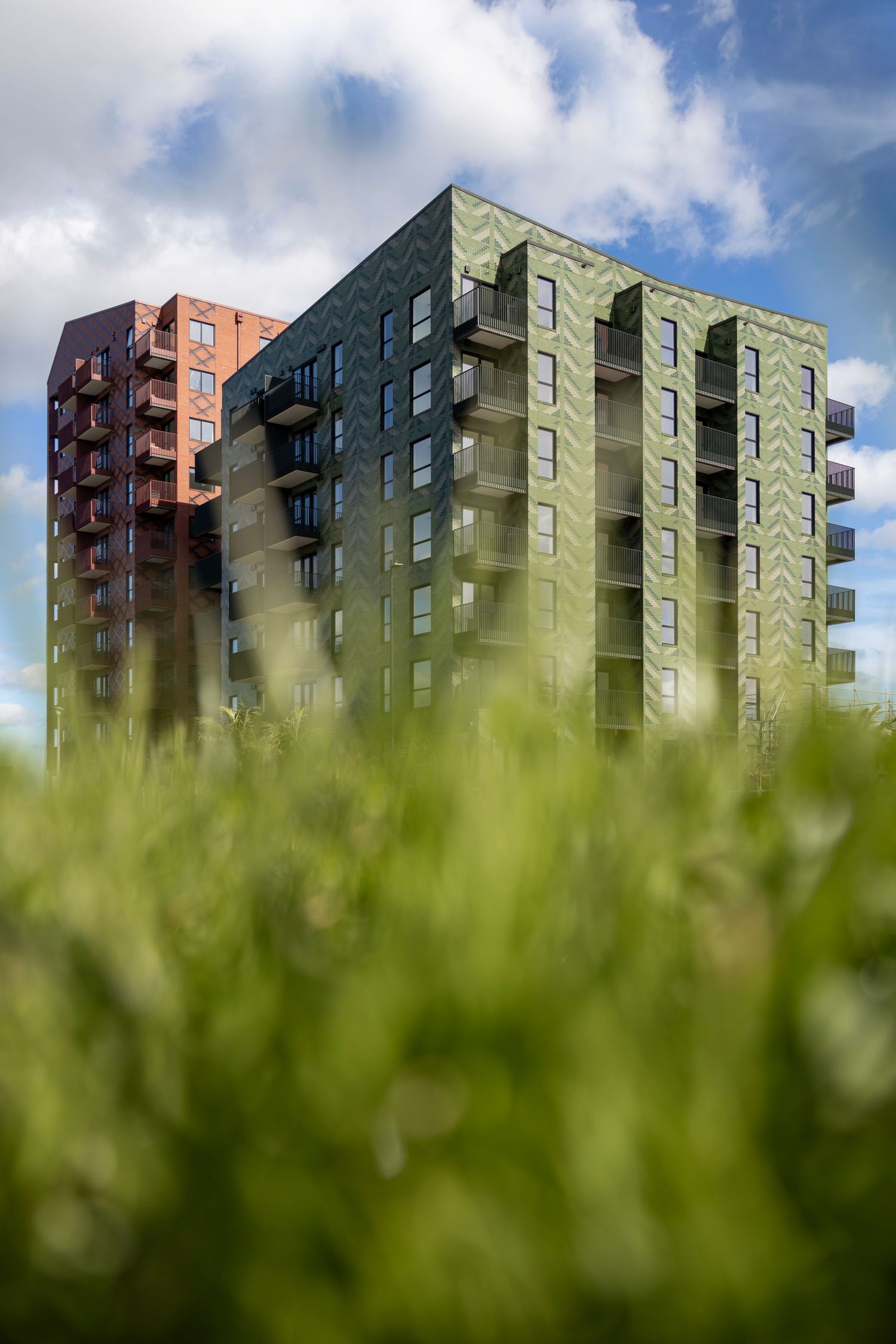 Twin green and red apartment towers rise behind blurred foreground grass under a blue sky.