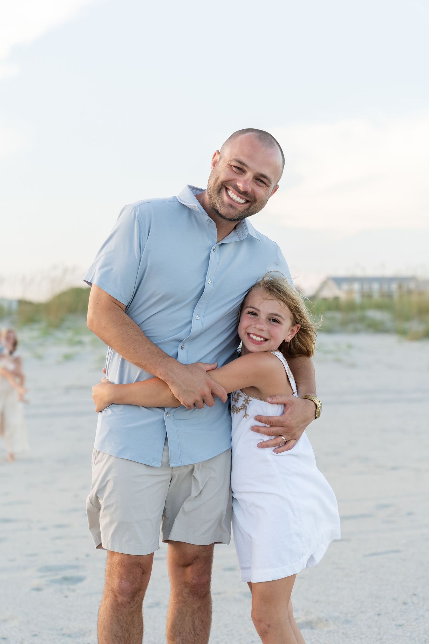 A father and child share a joyful embrace while wearing coordinated blue and white outfits on a sandy beach at sunset.