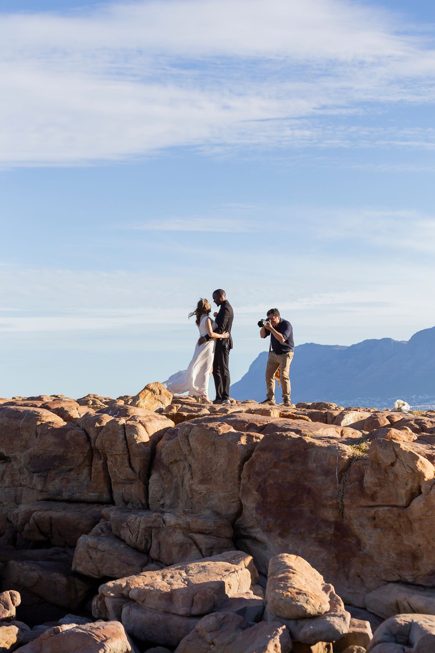 A photographer captures a couple on a rocky outcrop against mountain silhouettes and blue sky in the background.