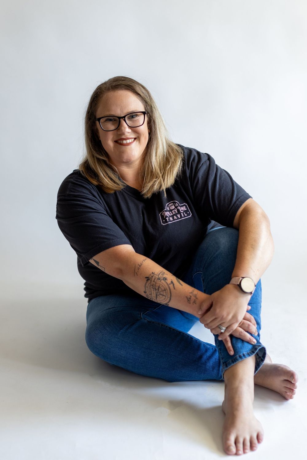 A person wearing glasses and a navy t-shirt sits cross-legged on a white background, displaying arm tattoos and a warm smile.