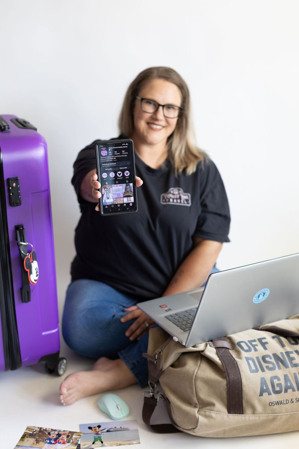 Person in glasses sits with laptop and purple luggage, displaying mobile phone screen while wearing company polo shirt.