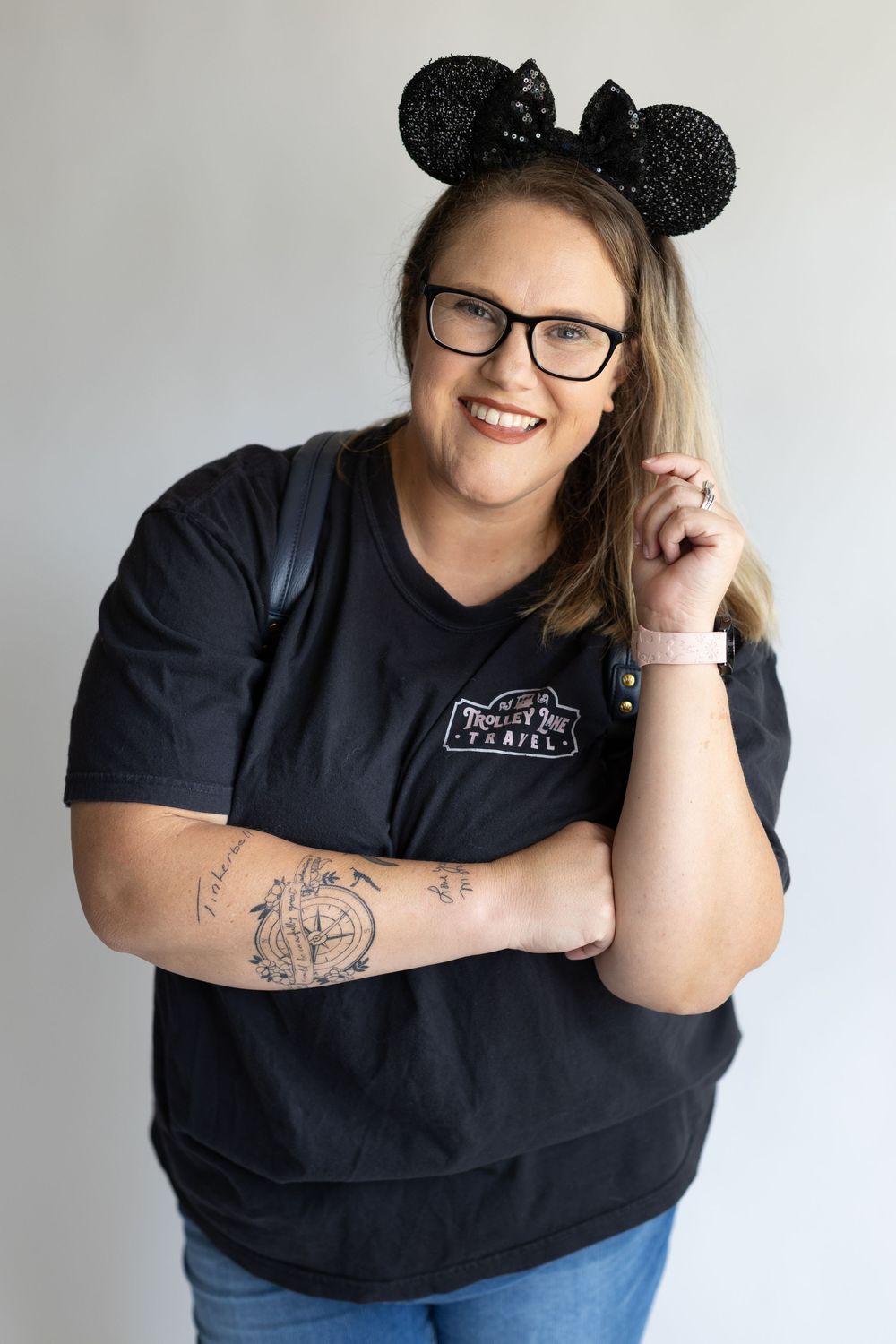 A person in glasses wearing a black t-shirt and sparkly Minnie Mouse ears poses against a light background with a tattoo on their arm.