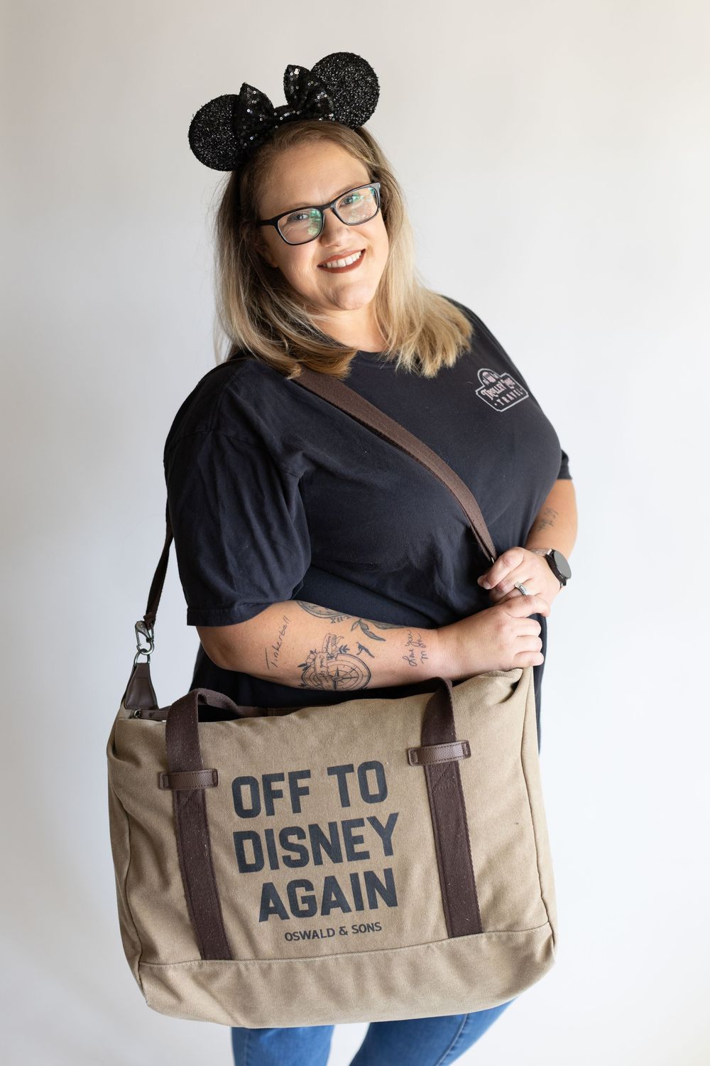 Person wearing Mickey Mouse ears poses with a tan canvas tote bag that says 'Off to Disney Again' against white background.