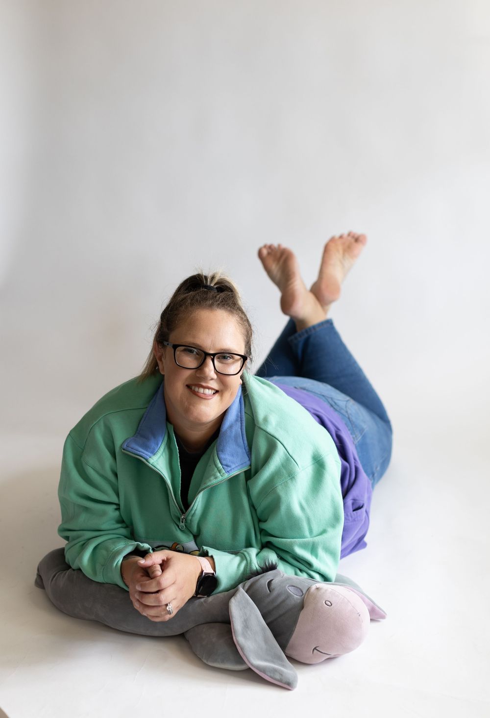 Person wearing glasses and a mint green jacket relaxes on floor in casual pose against white background.