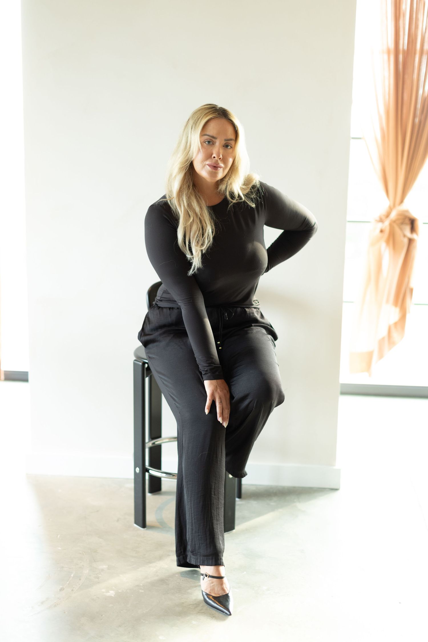 Woman in all black outfit with long blonde hair poses on bar stool in modern minimalist setting.