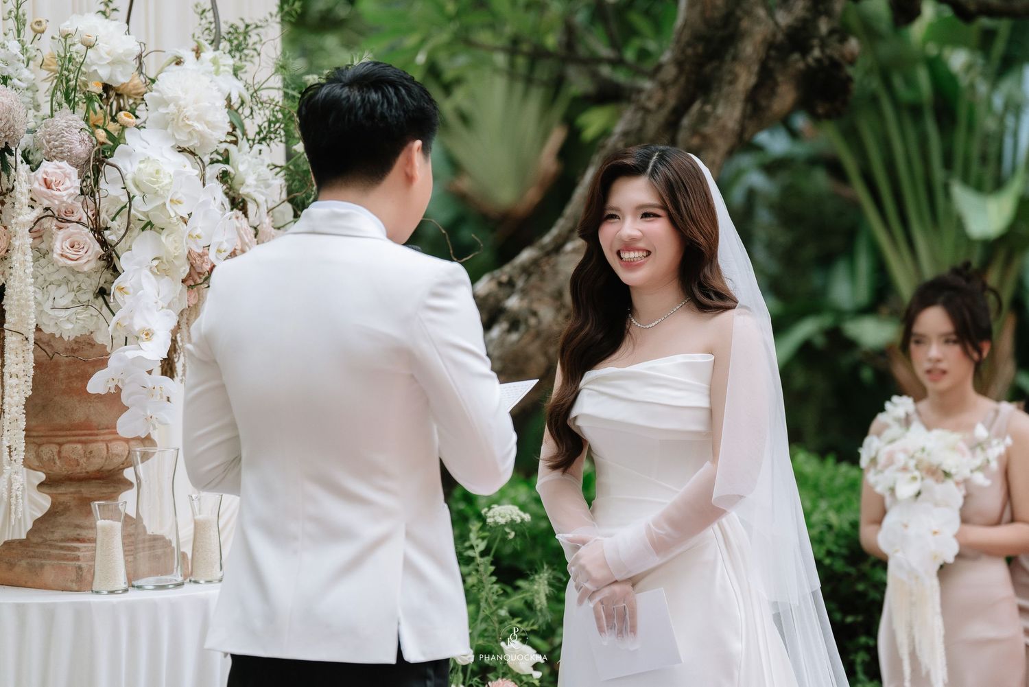 Smiling bride in white gown and veil listens to groom during outdoor wedding vow exchange ceremony.