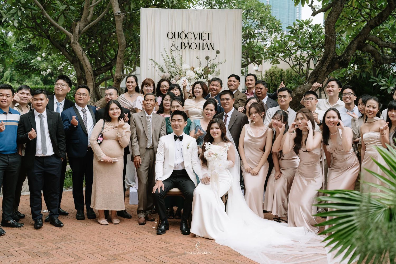 Wedding couple seated with large group of guests in formal attire outdoors at garden venue.
