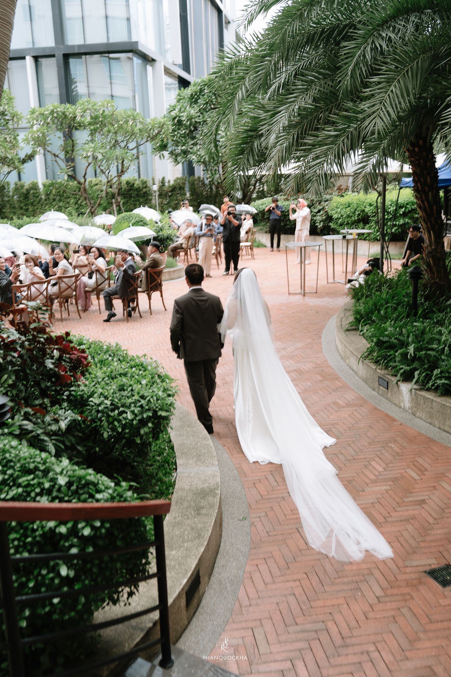 Bride with long flowing veil walks with groom along brick pathway toward wedding guests at garden ceremony.