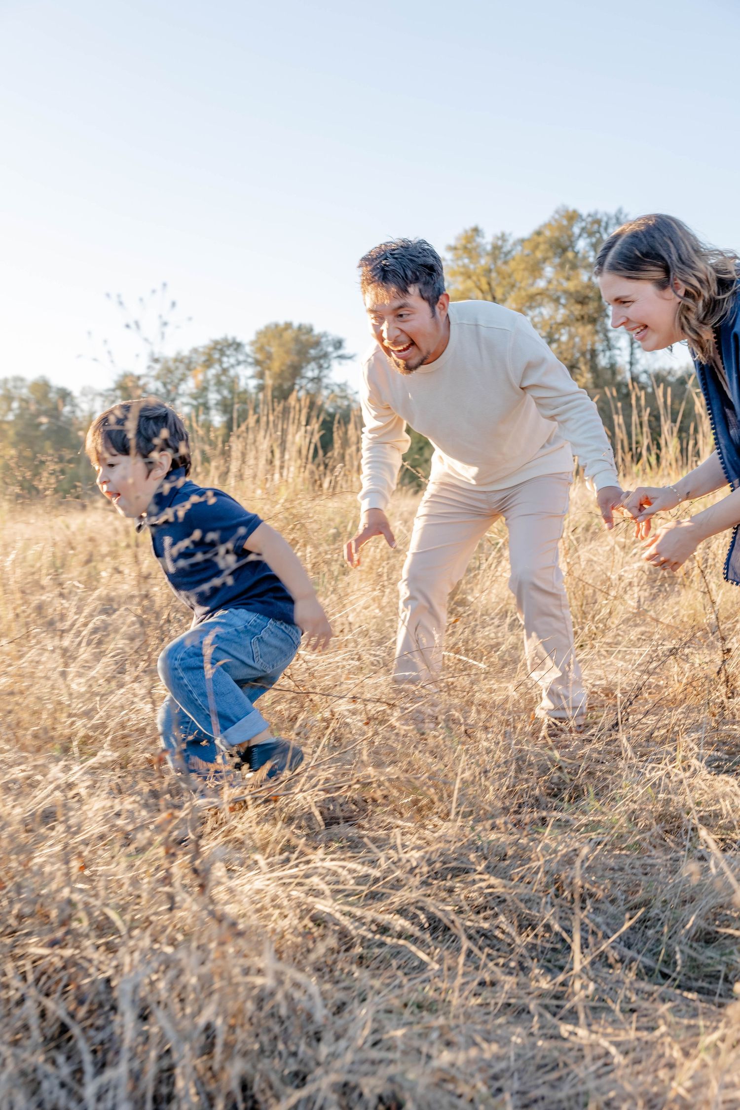 Family of three playing in golden field, parents chasing laughing child through dry grass on sunny day.
