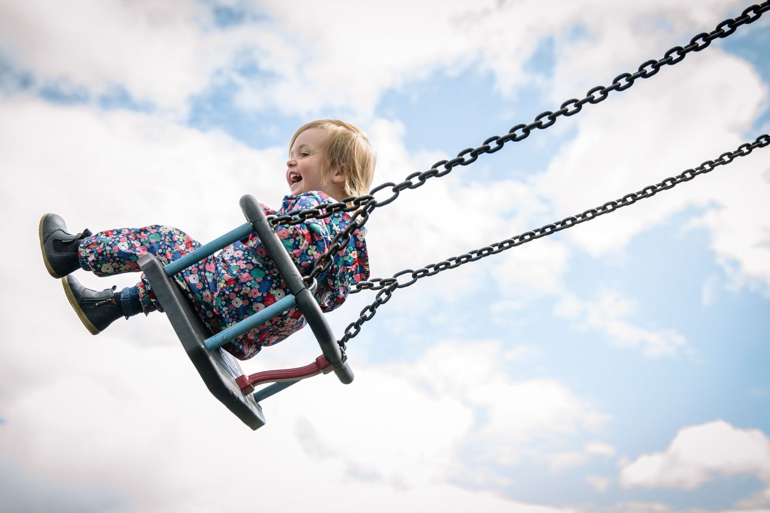Child swinging high in the air on a playground swing set against cloudy blue sky.