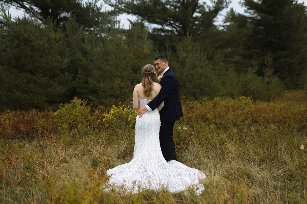 A romantic couple in wedding attire share an intimate moment in a rustic field surrounded by pine trees.