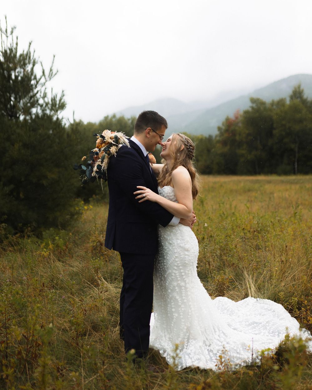 A romantic wedding couple embraces in a misty mountain meadow with lush greenery and a dramatic train on the wedding dress.