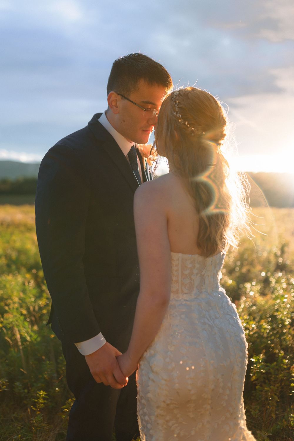 Newlywed couple shares intimate moment in golden field at sunset with sun rays creating ethereal glow around them.
