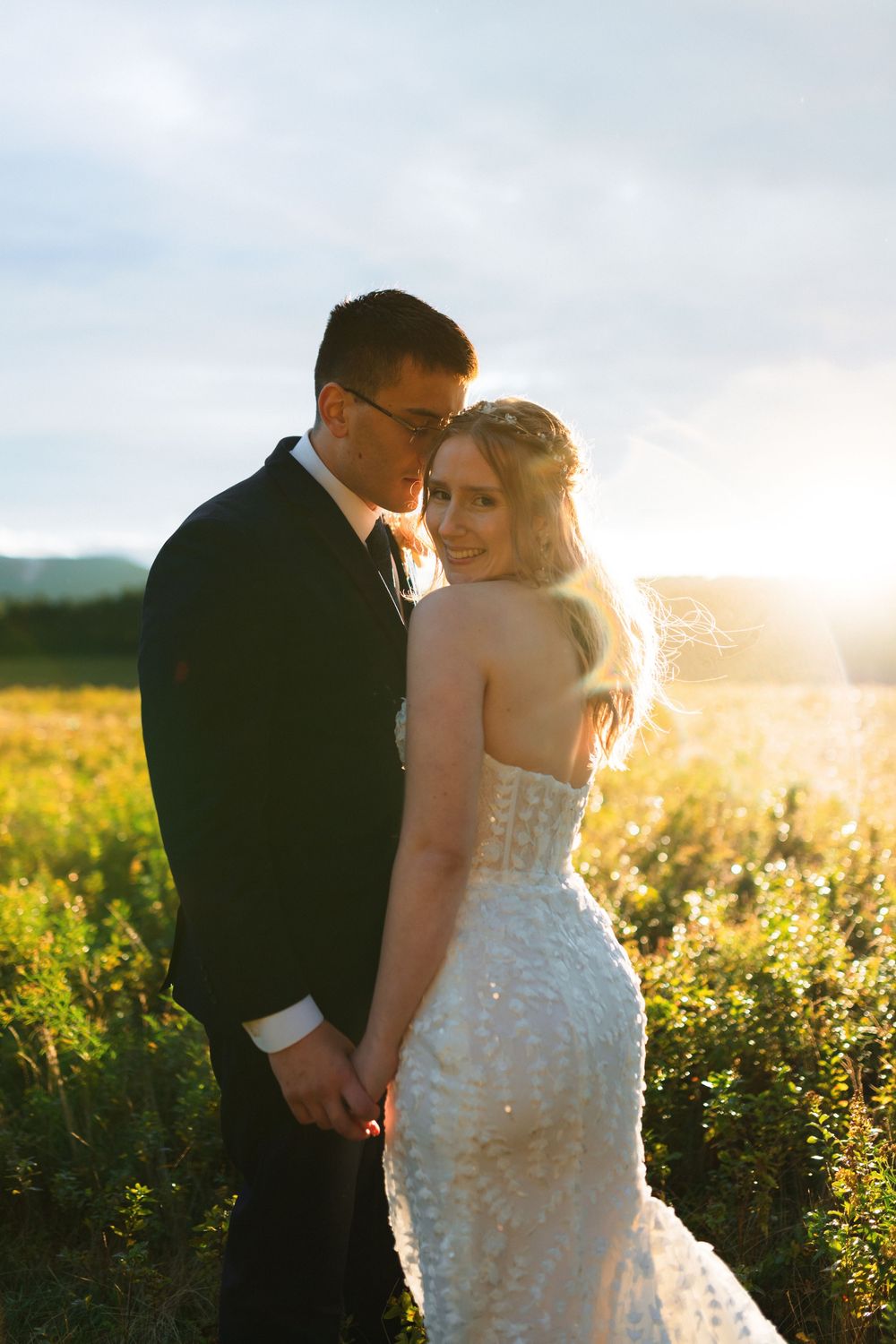 Romantic wedding couple embraces in golden sunlit meadow during sunset with bride wearing lace dress.