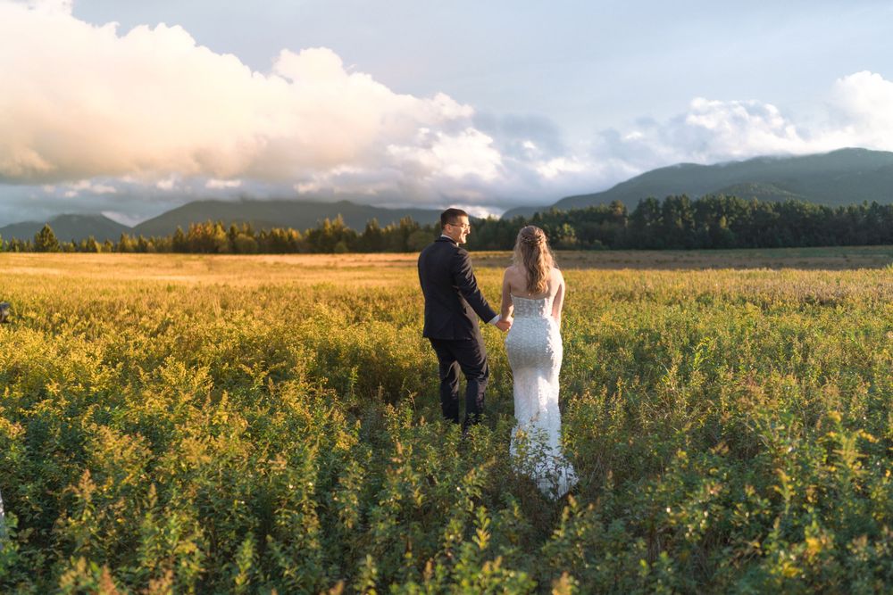 A couple stands in a golden wildflower field at sunset with majestic mountain peaks in the background.