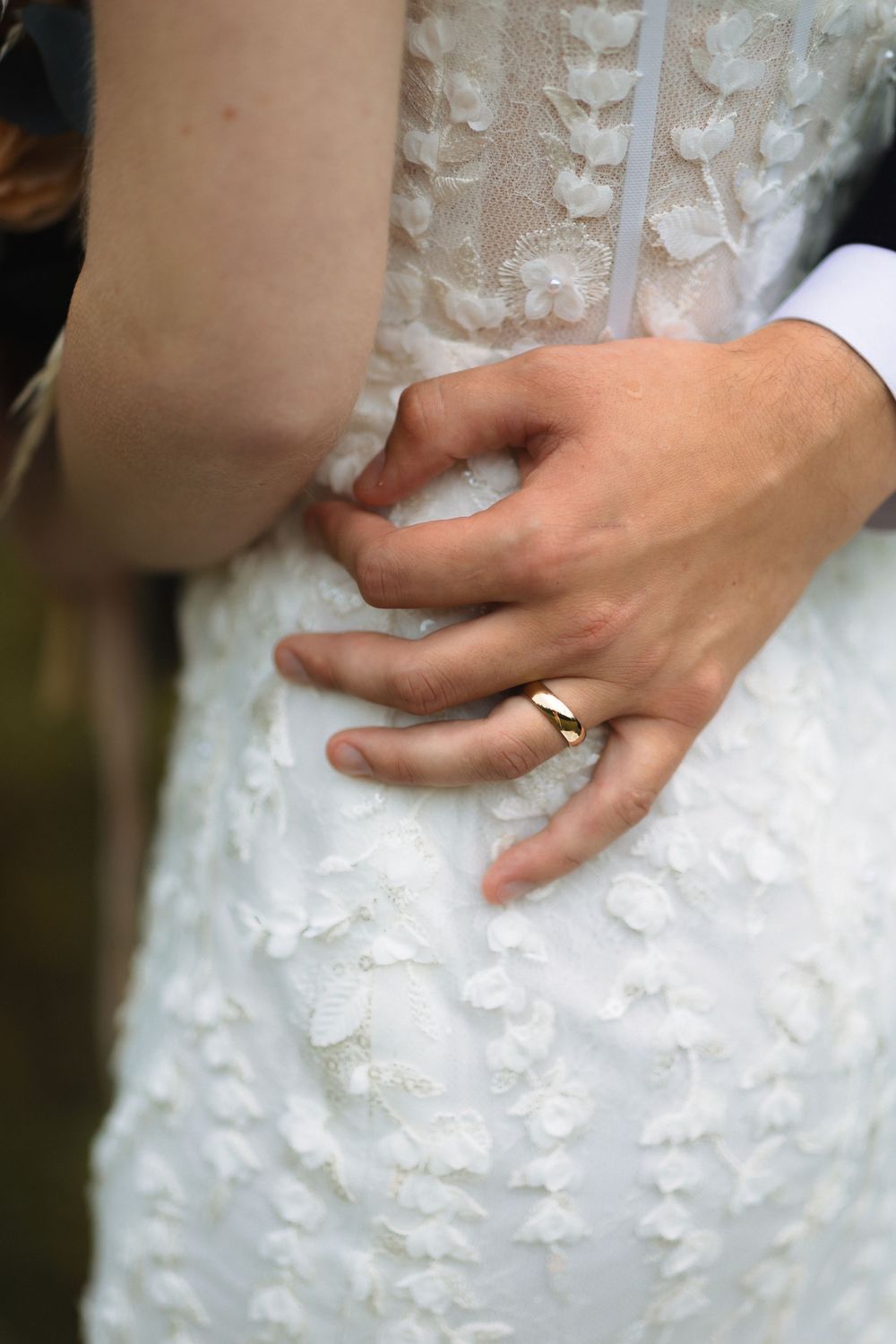 Close up of hands on a white lace wedding dress with a gold wedding band visible on finger.