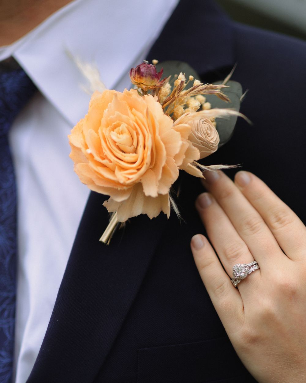 A peach-colored rose boutonniere pinned to a navy suit lapel next to a hand wearing an elegant engagement ring.