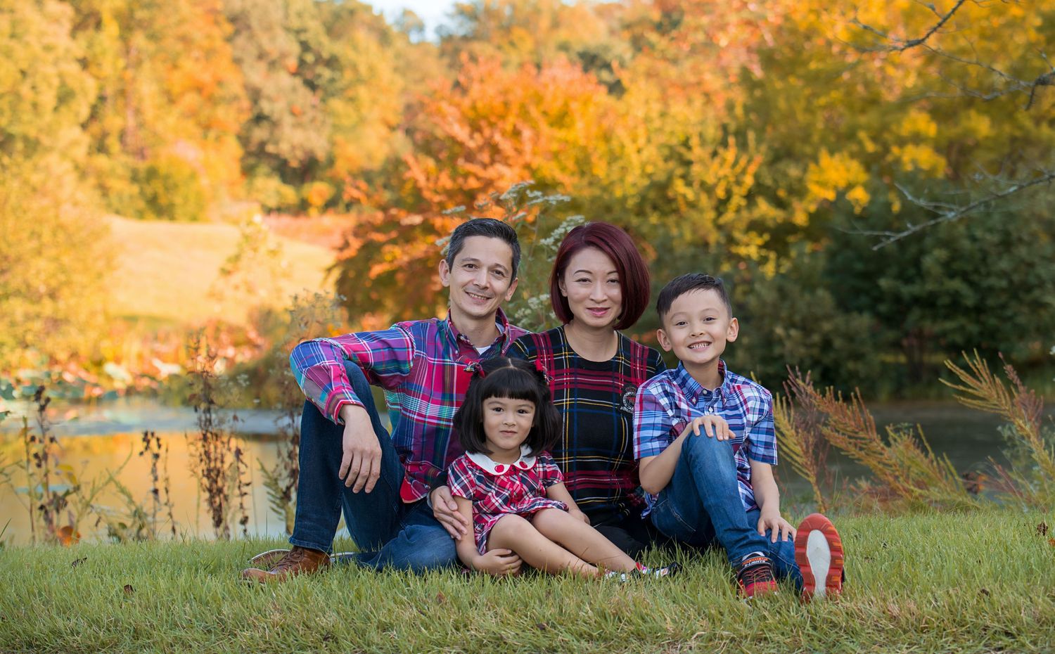 Family sitting together on grass during autumn with colorful fall foliage in background.