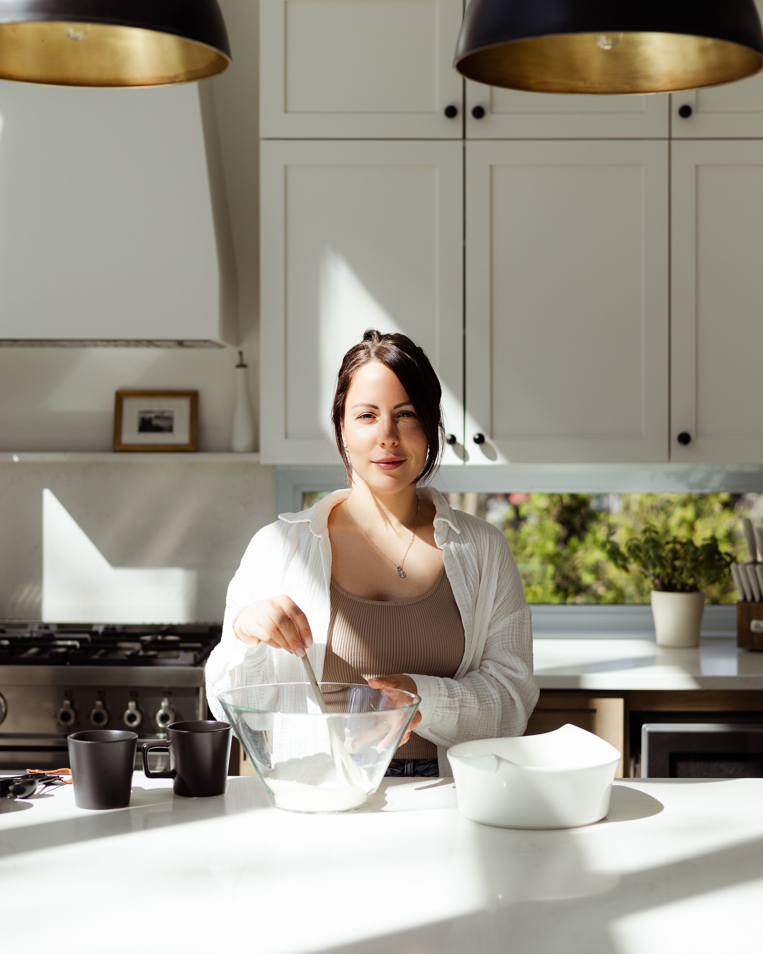 Person in white robe mixing ingredients in sunlit modern kitchen with pendant lights.