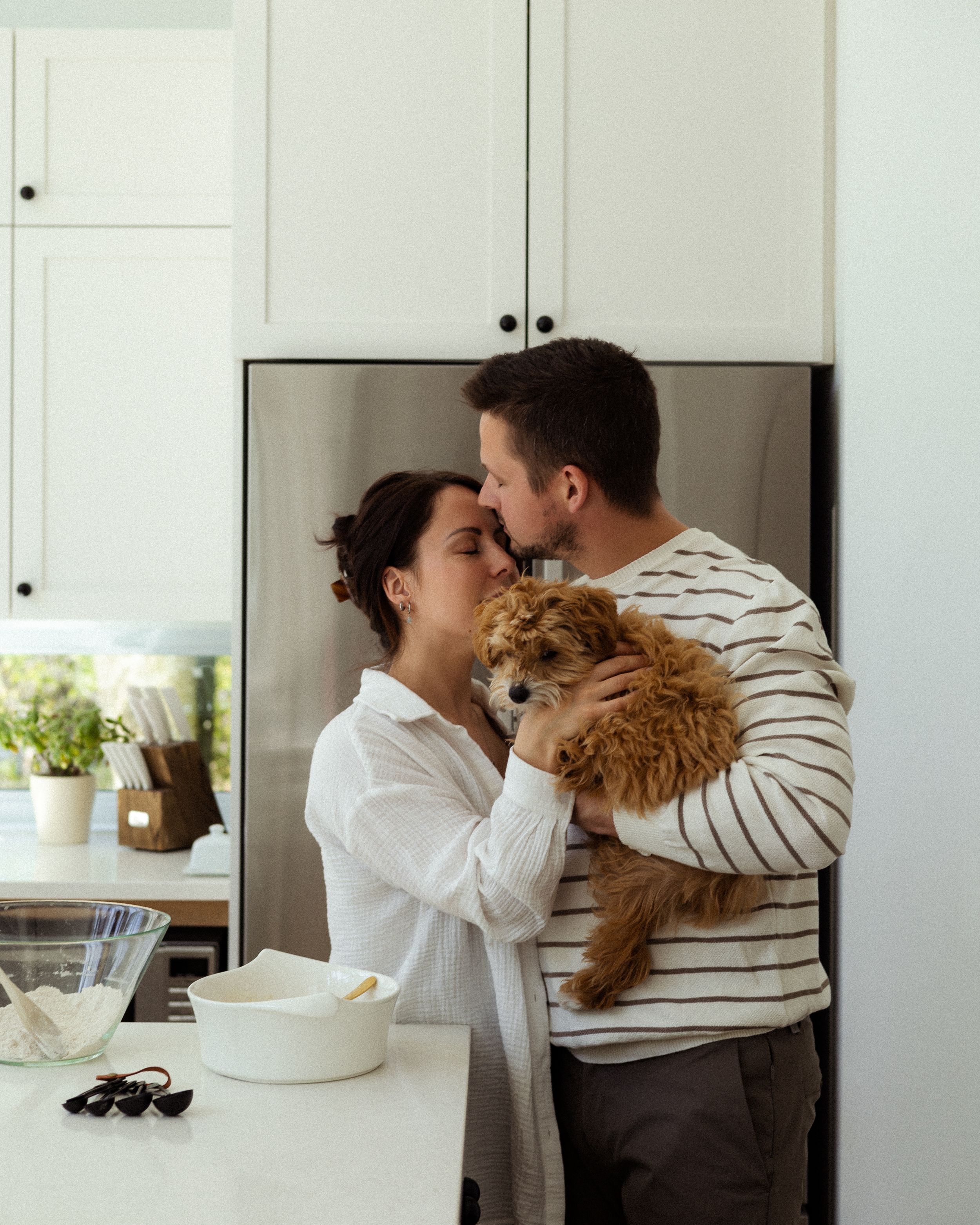 In bright kitchen, couple holds brown dog while sharing intimate moment.
