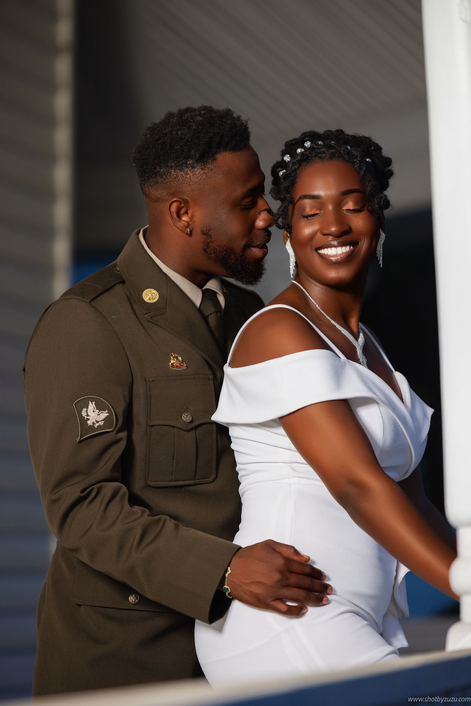 Military groom in dress uniform embraces bride in elegant white off-shoulder wedding gown during romantic photoshoot.