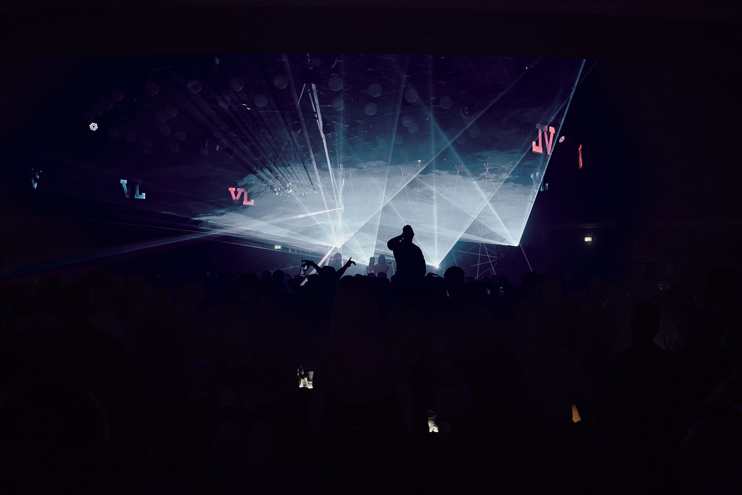 Concert venue with dramatic white light beams cutting through darkness above silhouetted crowd.