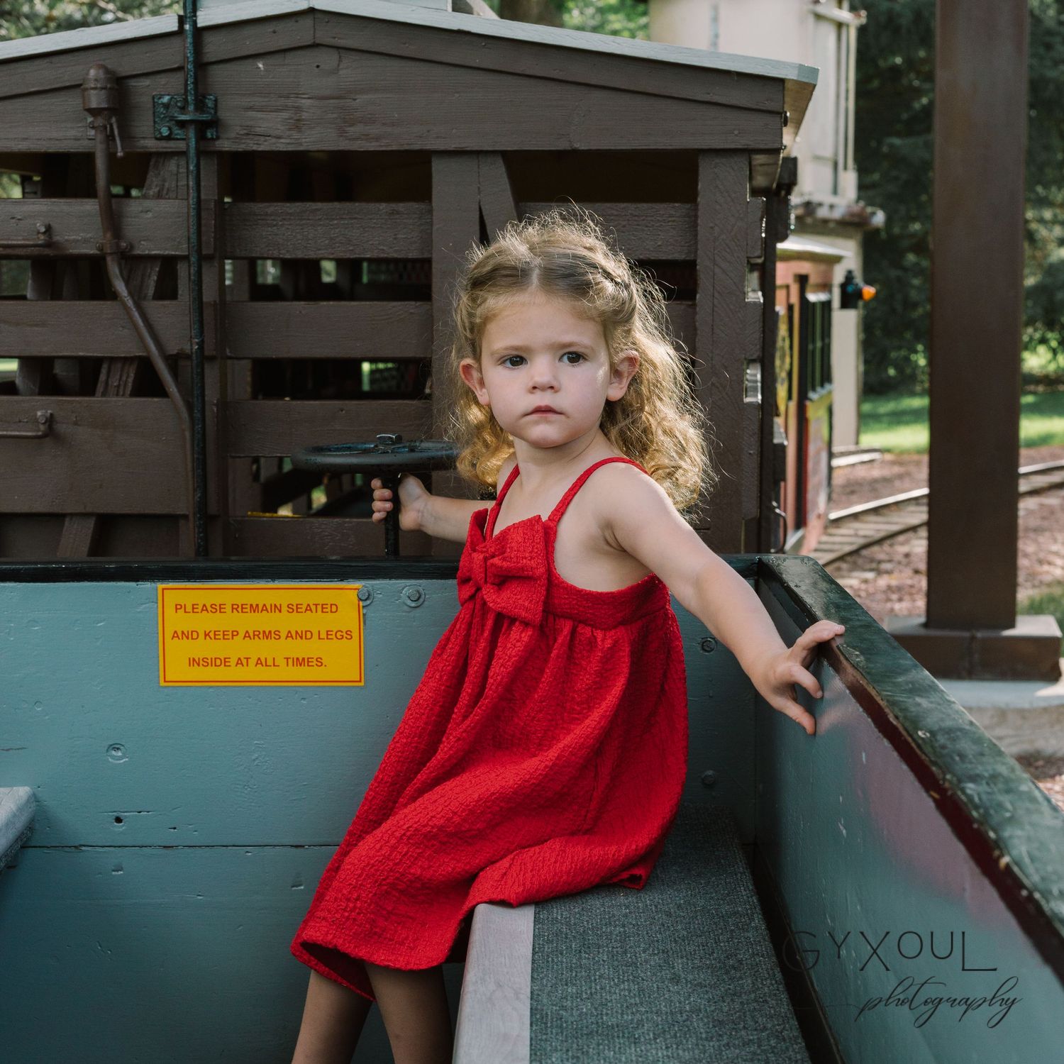 A child in a red dress leans against a wooden structure outdoors.