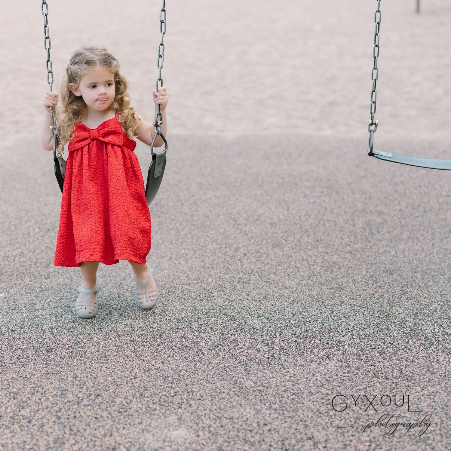 A child in a bright red dress stands holding playground swing chains.