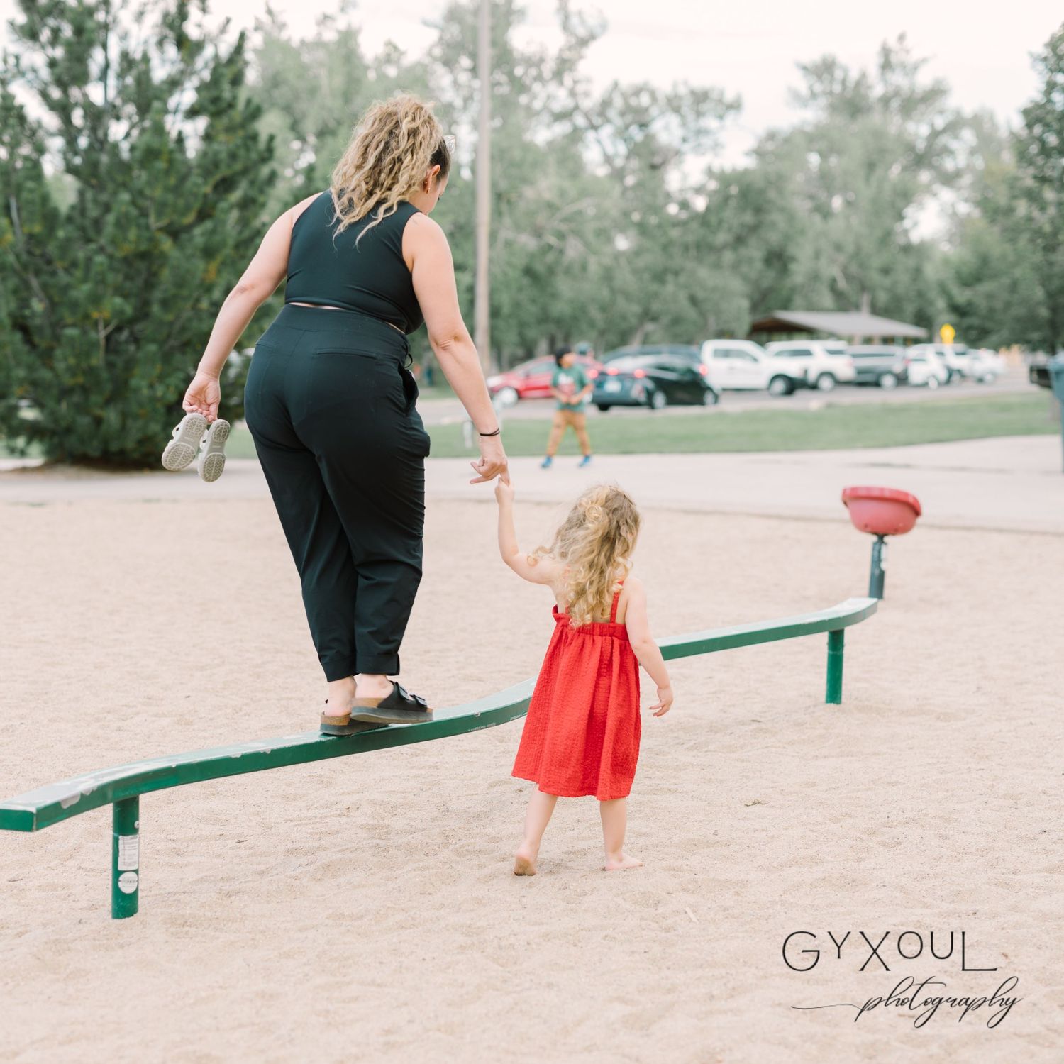 Two people balance together on a green playground balance beam in a park setting.