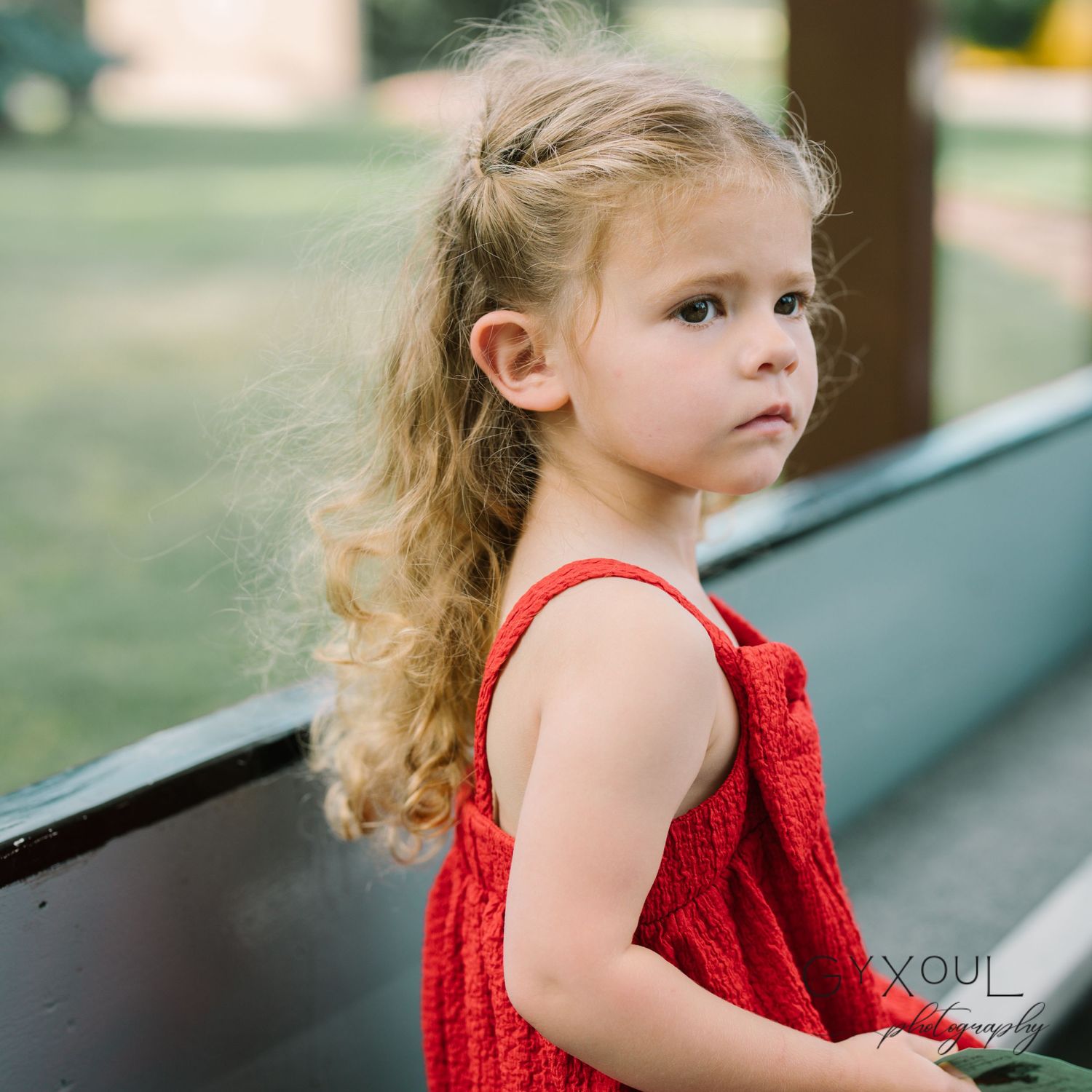 A child in a red dress stands thoughtfully near a wooden structure in an outdoor setting.